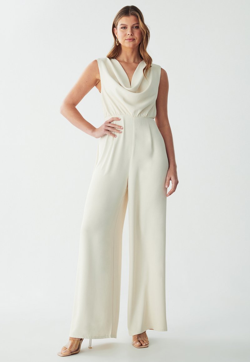 Combinaison couleur crème avec un décolleté drapé, sans manches, et une taille élastique. Silhouette à jambes larges et tissu lisse et fluide.