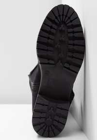 Semelle de botte noire avec des motifs de crampons profonds et la taille 37 indiquée, positionnée à la verticale contre une surface blanche.