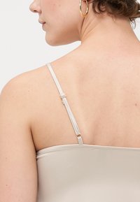 Beige cami med tynne, justerbare stropper. Partikkeltekstur og enkelt design, som viser et nærbilde av ryggen og skuldrene.