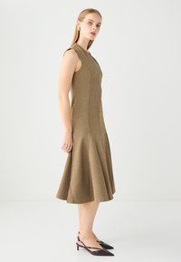Vestito marrone senza maniche con orlo svasato, caratterizzato da cuciture e una silhouette aderente. La modella indossa scarpe slingback nere con punta a mandorla.