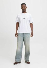 Camiseta blanca de algodón con logo bordado; vaqueros de denim azul claro de corte amplio con un acabado desgastado; zapatillas marrones con acentos blancos.