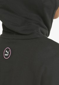 Hoodie noir avec une finition texturée, mettant en avant un logo circulaire Puma rose et noir au dos. Le tissu semble doux et léger.