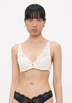 Camisola de renda em creme com aro, apresentando painéis de renda transparente e alças ajustáveis. Combinada com uma cueca de renda preta.