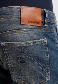 Blå denimjeans med detaljerad sömnad på bakfickan och en brun läderlogotyp med texten "CHASIN'" ovanför fickan.