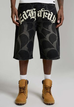 Uomo che indossa pantaloncini neri con scritta grafica "Death a Fady", calzini bianchi, stivali da lavoro color cuoio e una camicia bianca, in piedi davanti a uno sfondo grigio.