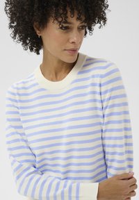 Femme aux cheveux bouclés portant un pull à rayures blanches et bleu clair, regardant sur le côté avec les bras légèrement croisés.