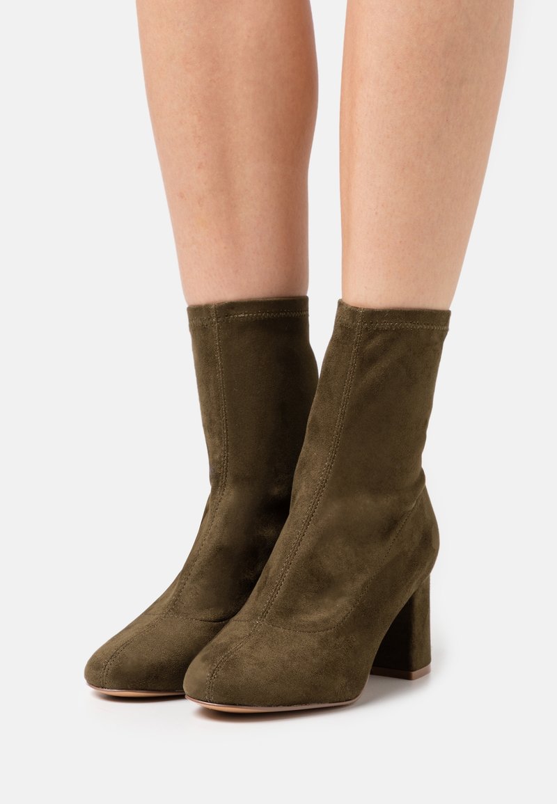 Bottines en daim marron à talons carrés portées sur des jambes nues sur un fond clair uni.