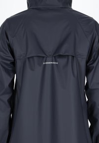 Granatowa kurtka przeciwdeszczowa z kapturem, wyposażona w guziki w tylnej wentylacji, o gładkiej teksturze, z logo "SWEDEMOUNT" w białym kolorze na dolnej części pleców.