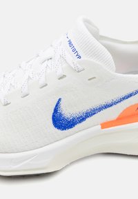 Witte sneaker met een gebreid bovenwerk, voorzien van een grote blauwe Nike swoosh, oranje accenten op de hiel en een gestructureerde zool voor grip.