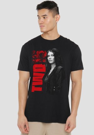 Young man wearing black TWD 5 graphic tee with female character portrait and red vertical text on front, beige pants, neutral background.