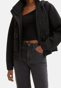 Giacca nera corta con cappuccio, chiusura a bottoni e zip, e due tasche frontali. Indossata sopra un top nero a coste e jeans grigio scuro.