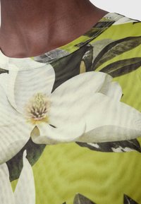 Blomster mesh top med en stor hvid magnolia mod en livlig grøn baggrund. Tekstiler med struktureret stof og rund halsudskæring.