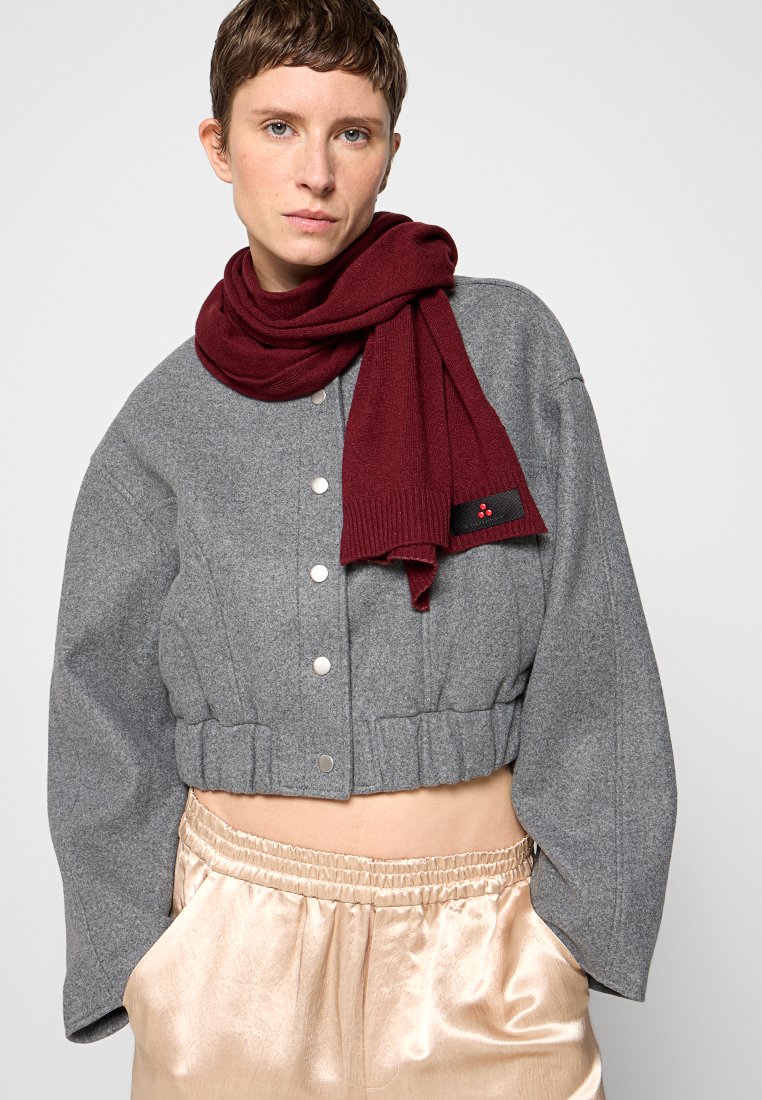 Person mit kurzen Haaren trägt eine graue cropped Jacke und einen burgunderfarbenen Schal, kombiniert mit glänzenden, hellen Hosen. Neutraler Hintergrund.