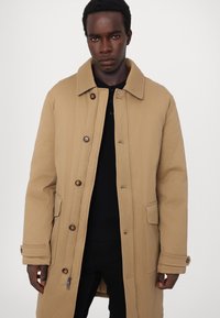Beige long coat made from fabric with a smooth texture, featuring a collar, button closure, side pockets, and a zipper detail at the hem.