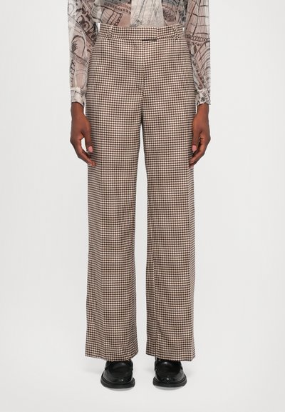 Weit geschnittene Hose mit einem braun-weißen Hahnentrittmuster, glatter Textur und tailliertem Bund. Darunter werden schwarze Schuhe getragen.