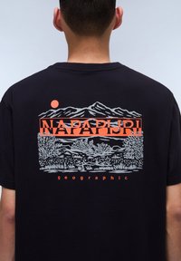 T-shirt noir avec un motif graphique de montagnes et un soleil en orange et gris. Le texte indique "NAPAPIJRI" en haut et "geographic" en bas.