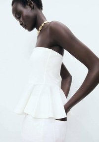 White strapless peplum top with a fitted bodice, textured fabric, and a flared hem. The model wears a gold necklace, showcasing an elegant look.