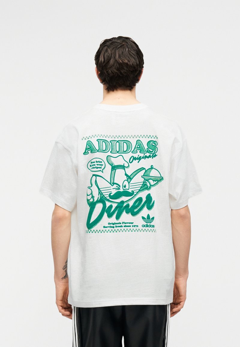 Personne portant un t-shirt blanc Adidas avec un graphisme rétro vert de diner montrant une banane chef faisant un clin d'œil et tenant un plateau de service.