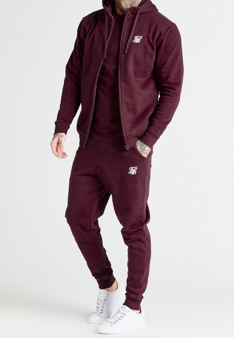 Homme portant un sweat à capuche zippé bordeaux assorti et un pantalon de jogging avec un logo blanc, des baskets blanches, des tatouages visibles sur les mains et le cou.