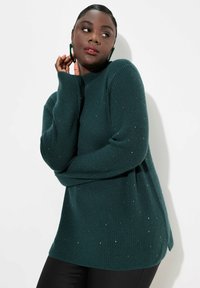 Teal stickad tröja med hög krage, långa ärmar och sidöppningar. Har metalliska detaljer spridda över hela plagget. Bärs med svarta byxor.