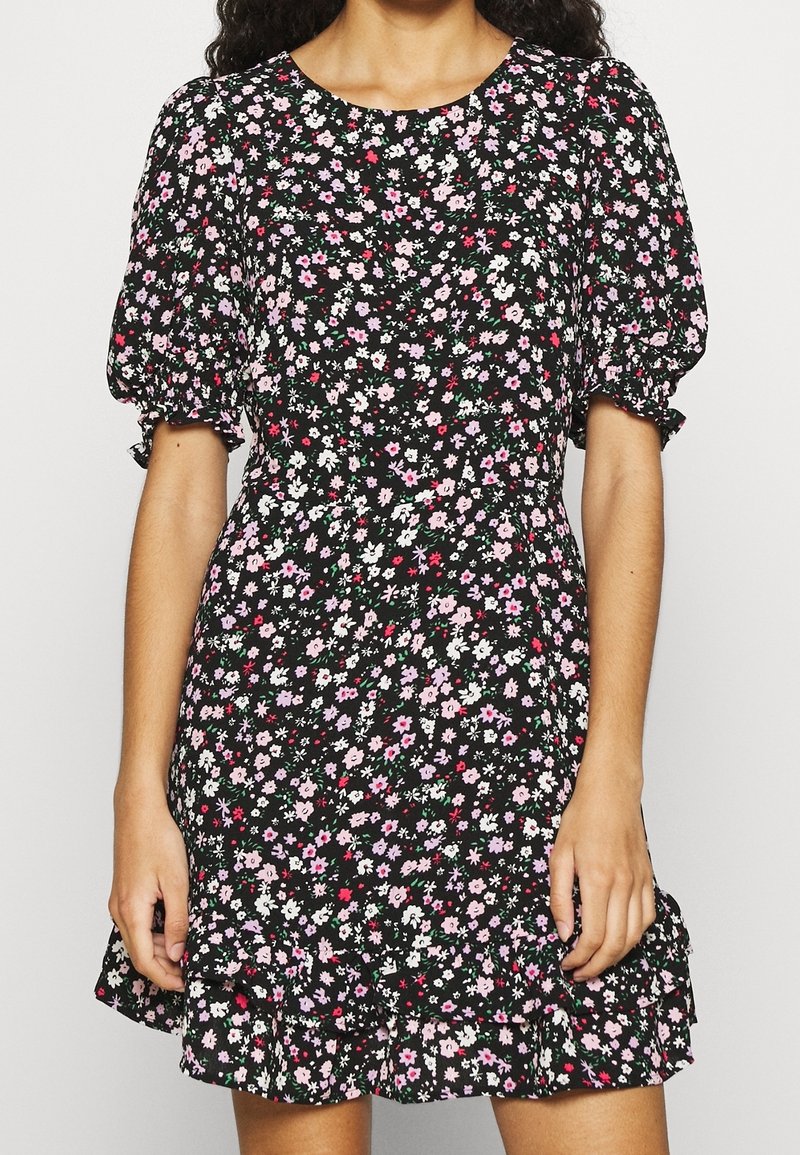 Femme portant une robe noire à fleurs avec des petites fleurs roses, blanches et rouges, des manches courtes bouffantes et un ourlet à volants.