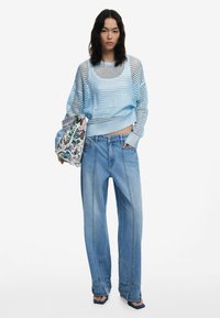 Helles blauer Netzpullover mit weiten Ärmeln, kombiniert mit hoch sitzenden, locker geschnittenen hellblauen Jeans. Das Model hält eine gemusterte Tasche.