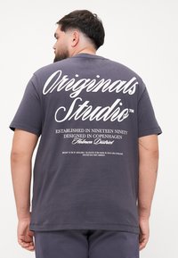 Tmavě šedé bavlněné tričko s bílým textem na zadn�í straně. Design obsahuje "Originals Studio" a další detaily v různých velikostech písma.