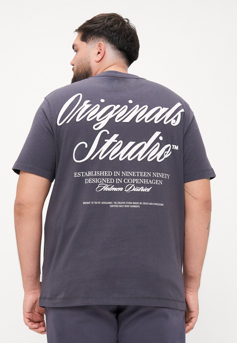 Tmavě šedé bavlněné tričko s bílým textem na zadní straně. Design obsahuje "Originals Studio" a další detaily v různých velikostech písma.