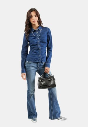 Vrouw draagt een gerimpeld blauw denimshirt, wijd uitlopende spijkerbroek, zilveren schoenen, houdt een zwarte gestructureerde handtas vast tegen een effen achtergrond.