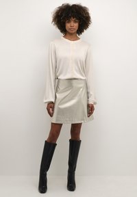 Világos bézs gombos ing hosszú ujjakkal, fémes ezüst mini szoknyával és térd fölé érő fekete csizmával párosítva. Sima textúra az egész felületen.