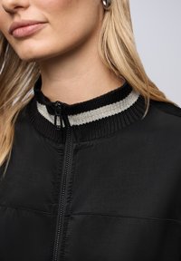 Veste zippée noire avec un col haut, ornée de rayures noires et grises alternées. Tissu lisse avec un design élégant et moderne.