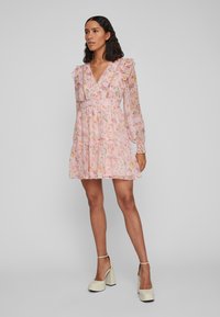 Blomstrete rosa kjole med lange ermer, volanger og dyp V-hals. Har et lagdelt skjørt med blonde detaljer. Brukt med beige platåheels.
