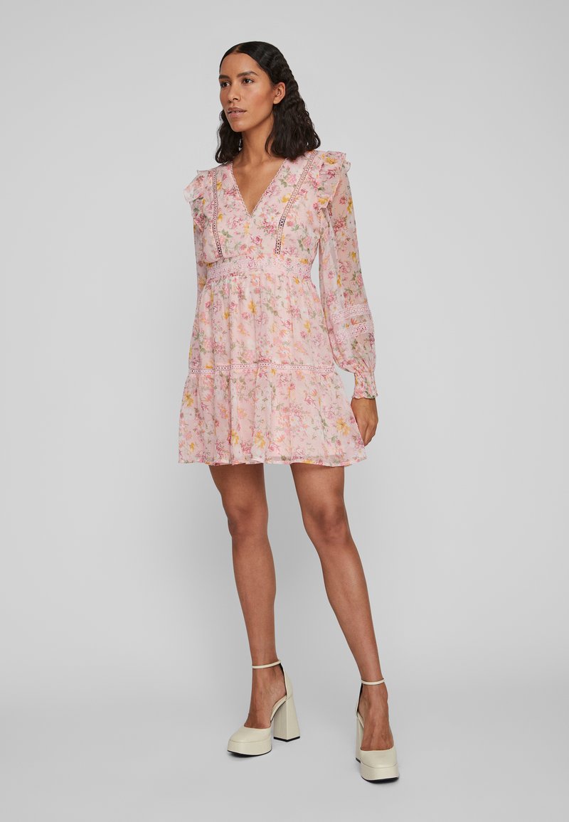 Blomstrete rosa kjole med lange ermer, volanger og dyp V-hals. Har et lagdelt skjørt med blonde detaljer. Brukt med beige platåheels.