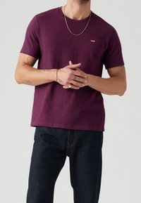 T-shirt en coton violet à manches courtes avec un col côtelé et un petit logo rouge sur la poitrine, associé à un jean bleu foncé.