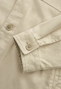 Beige színű pamut ingujj, hajtott mandzsettával, két gombbal és ferde varrási részletekkel. Simai textúra, amely diszkrét csíkozást mutat.