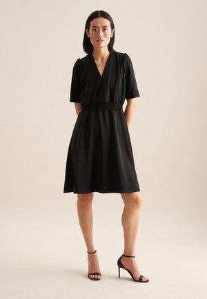 Robe noire avec manches courtes, décolleté en V et taille smockée. Tissu fluide tombant jusqu'à mi-cuisse. Associée à des sandales à talons hauts noires.