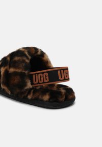 Chausson ouvert à l’avant avec imprimé léopard et semelle noire, avec une sangle élastique marron et noire avec le mot "UGG" répété.