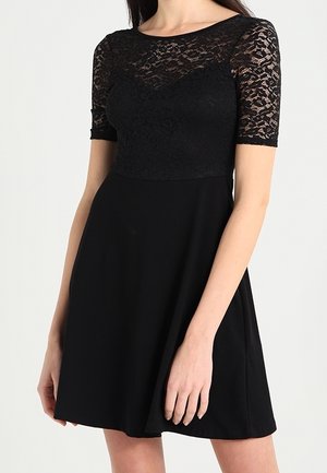Femme portant une robe noire longueur genoux avec un haut en dentelle, des manches courtes et une taille cintrée s'évasant en jupe trapèze.