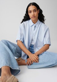 Vrouw met lang krullend haar gezeten in kleermakerszit, gekleed in een lichtblauw overhemd met korte mouwen, een wit shirt, wijd uitlopende spijkerbroek en beige schoenen.