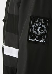 Černá bunda s vodorovnými černými a bílými pruhy. Na levém rukávu se nachází velká černá nášivka s bílým logem. Hladká textura.