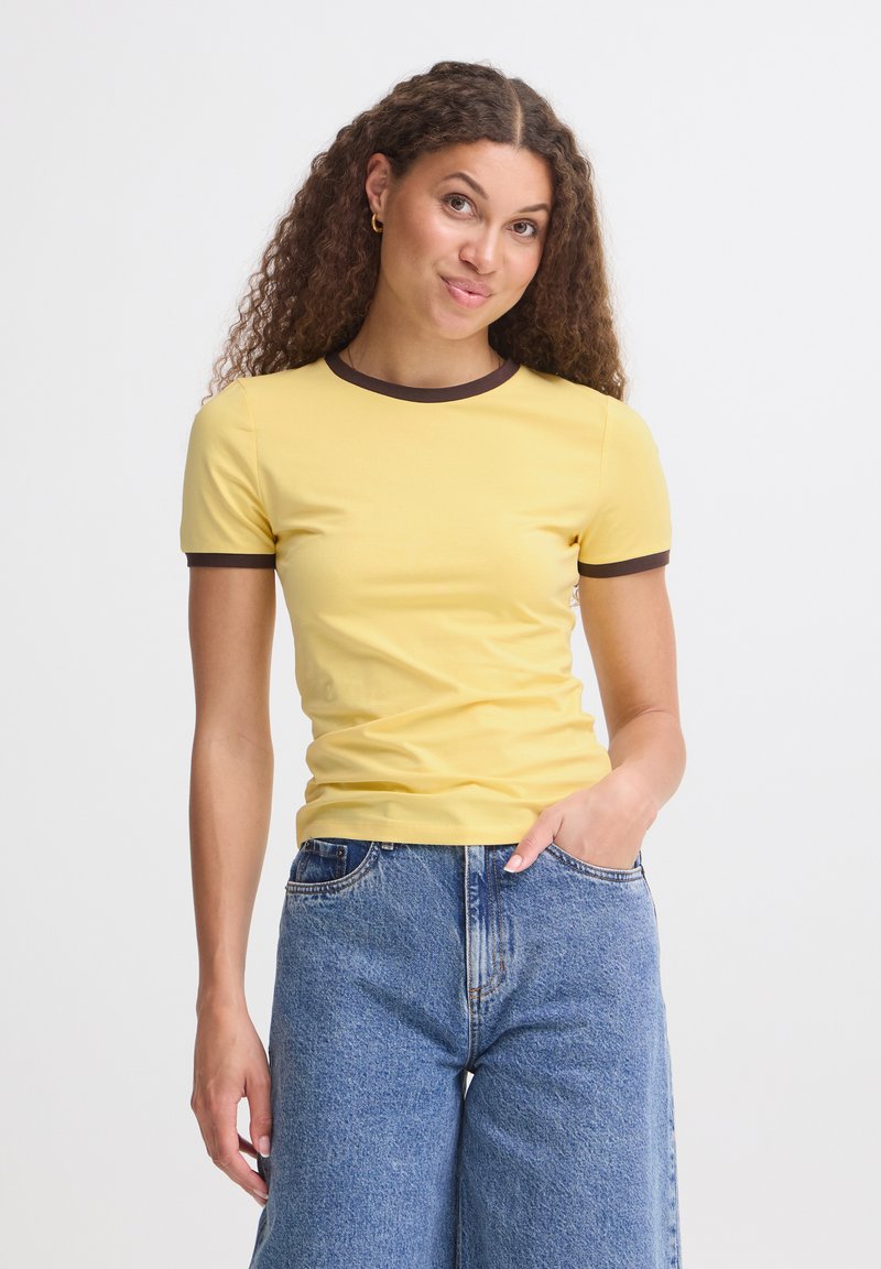 Femme aux cheveux bouclés portant un t-shirt jaune avec une bordure foncée et un jean bleu, debout avec une main dans sa poche sur un fond blanc.