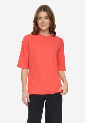 Jeune femme portant un T-shirt corail à manches courtes et oversize ainsi qu'un pantalon noir, debout devant un fond blanc, souriant légèrement.