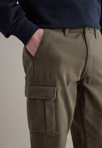 Mann trägt olivgrüne Cargohosen, steckt die Hand in die Tasche und trägt ein marineblaues Langarmshirt vor einem schlichten Hintergrund.