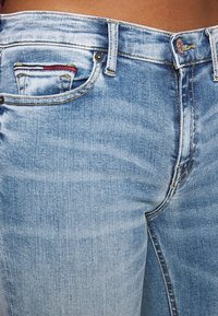 Jeans de denim azul claro con un lavado desgastado; cuenta con un pequeño bolsillo frontal con un acento a rayas, costuras visibles y un cierre de botón metálico.