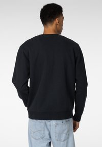 Svart genser med lange ermer, rund hals, og ribbede mansjetter. Merkbar søm detaljer ved kanten, brukt med lyseblå jeans.