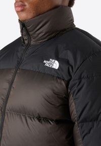 Přifukovací bunda s černou horní částí a tmavě hnědou spodní částí, má vysoký límec a logo The North Face na hrudi.
