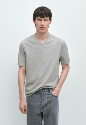 Giovane uomo con capelli corti e scuri, che indossa una T-shirt grigia a scollo a V e jeans grigi, in piedi davanti a uno sfondo chiaro e semplice.