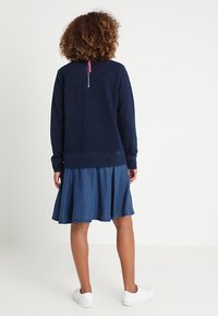 Marineblauer Sweatshirt mit einem Reißverschluss auf der Rückseite, kombiniert mit einem Jeansrock. Beides ist lässig, mit lockerer Passform und weichen Materialien. Weiße Sneakers vervollständigen den Look.
