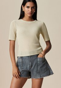 Kurzärmliges, figurbetontes, hellgelbes Oberteil aus einem strukturierten, schimmernden Stoff, kombiniert mit hoch taillierten, dunklen Denim-Shorts mit großen Taschen.