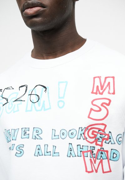 Κοντινό πλάνο ενός ατόμου που φοράει λευκό πουκάμισο με πολύχρωμο κείμενο που λέει «ΜΗΝ ΚΟΙΤΑΣ ΠΙΣΩ, ΟΛΑ ΕΙΝΑΙ ΜΠΡΟΣΤΑ» και κόκκινα κατακόρυφα στοιχισμένα γράμματα «MSGM».